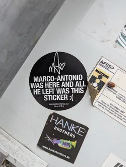 Street sticker Circular black sticker with white text and a stylized signature monogram at the top. The main text reads 'MARCO-ANTONIO WAS HERE AND ALL HE LEFT WAS THIS STICKER :('. Bottom text includes a social media handle.