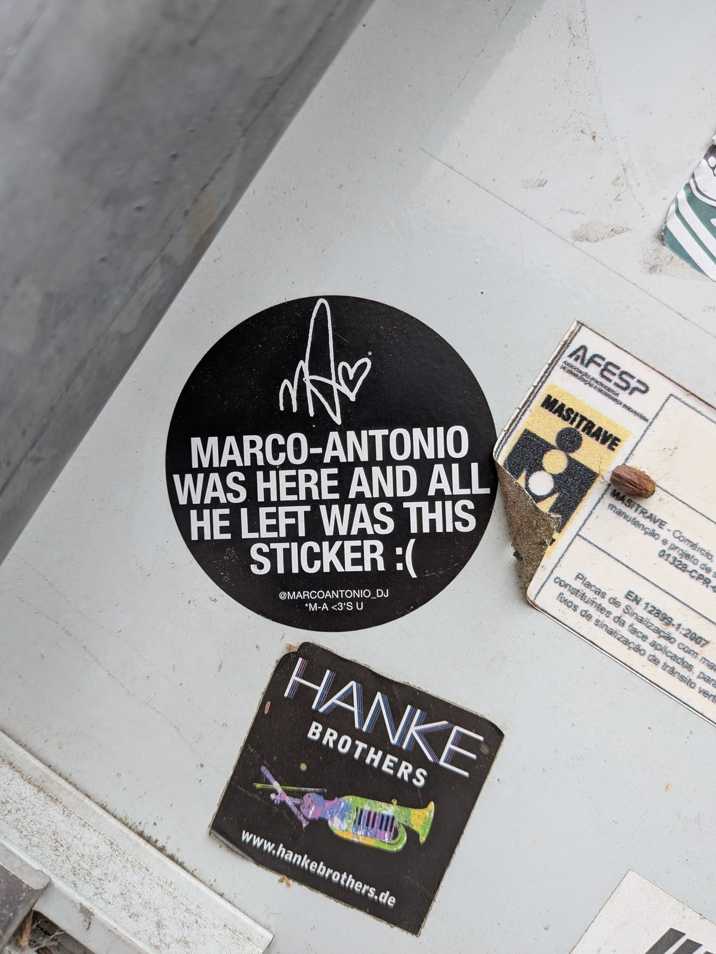 Circular black sticker with white text and a stylized signature monogram at the top. The main text reads 'MARCO-ANTONIO WAS HERE AND ALL HE LEFT WAS THIS STICKER :('. Bottom text includes a social media handle.