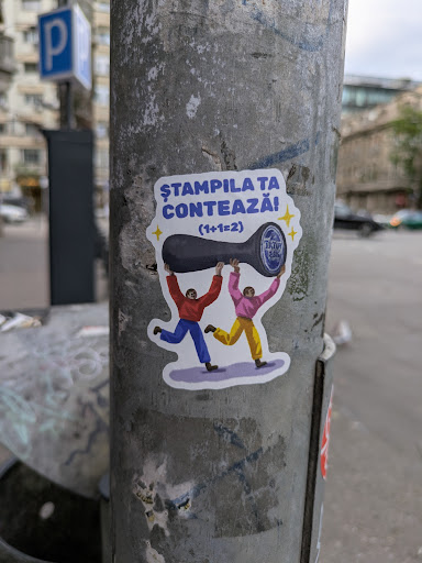 A sticker on a metal pole features two cartoon people celebrating. They are raising a stamp bearing text above their heads. The sticker also contains the text "Ștampila ta contează! (1+1=2)" written in blue.