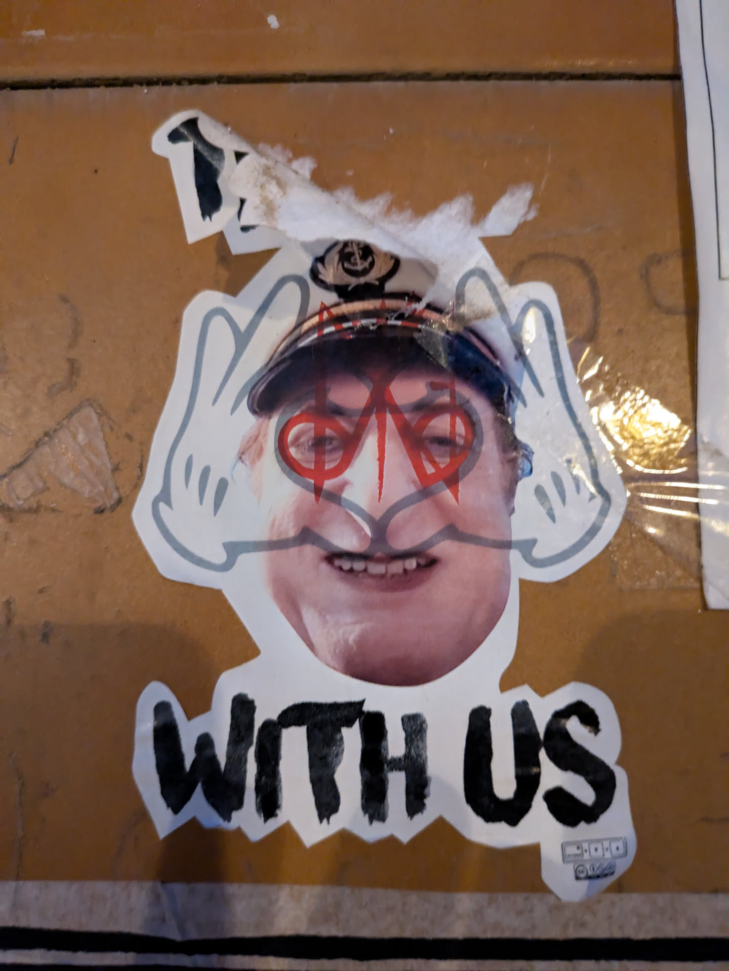 The sticker features a person's face with red circles drawn over their eyes, giving them an otherworldly appearance. They are wearing a hat, and Mickey Mouse-style hands flank their head. The words "WITH US" are printed below the image. A small, illegible logo is visible at the bottom right.