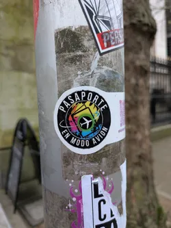 Street sticker A circular sticker with a white outer border. The central design features a stylized, open passport with pages rendered in a vibrant, abstract spectrum of colors including yellow, orange, red, pink, blue, and green. A white airplane silhouette is centrally placed, flying over a green circle. The words "PASAPORTE" are written in an arc at the top, and "EN MODO AVION" are written in an arc at the bottom, both in white capital letters against a black background that frames the colorful passport design.