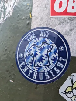 Street sticker The sticker is circular and predominantly blue with a white outer border. The text "THE KIT ENTHUSIAST" is written in white capital letters, curving around the perimeter. In the center, there is an outline of a t-shirt or jersey, filled with a repeating blue and white geometric pattern that resembles chevrons or pixels. Small white soccer ball icons are placed on either side of the t-shirt graphic, between the words "KIT" and "ENTHUSIAST" and between "THE" and "ENTHUSIAST" (though the latter is partially obscured). The overall design suggests a passion for sports jerseys or football kits.