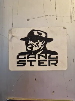 Street sticker A square sticker featuring a stylized illustration of a gangster in a fedora. The word 'GANGSTER' is written in a bold, block-like font beneath the illustration. The sticker has a worn, slightly aged look. 