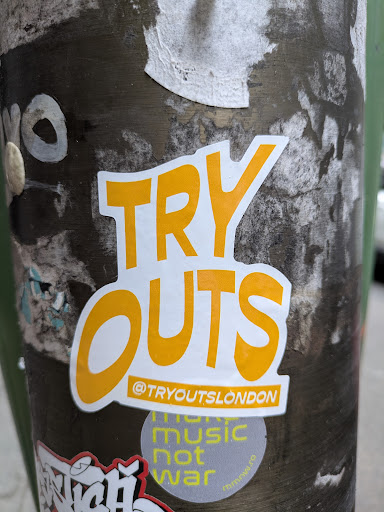 A 'TRY OUTS' sticker with an Instagram handle is pasted on a pole. The sticker has a white background and bright yellow text. The text has two levels, 'TRY' on top and 'OUTS' below it. Below the text, there is an Instagram handle. Underneath this sticker, there is another circular 'make music not war' sticker. The pole has some other stickers on it and is slightly weathered.