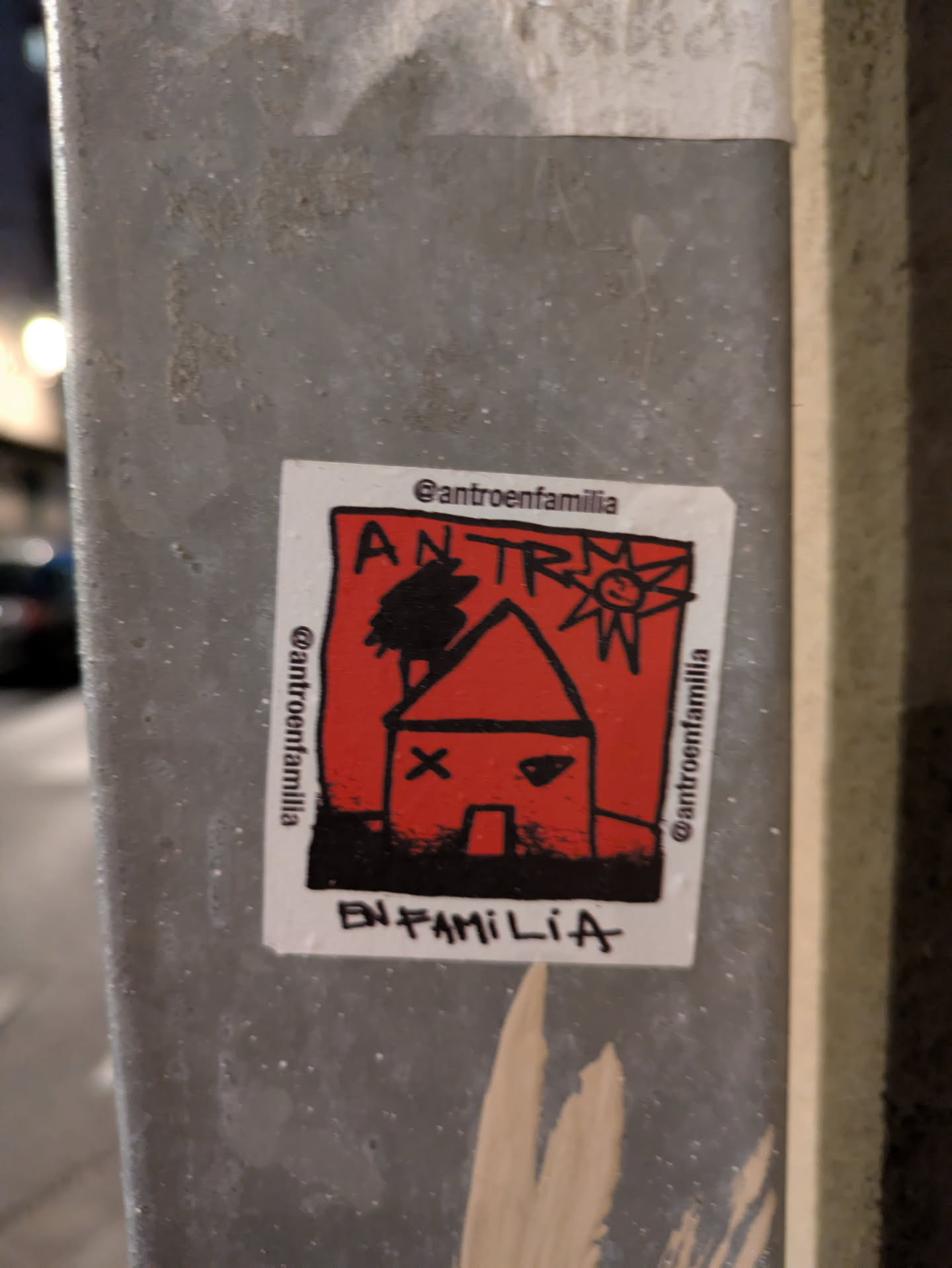 Street sticker This is a square sticker with a white border, showcasing a red house with black outlines and details. A black tree stands to the left of the house, and a sun with a smiley face is in the upper right corner. The words 'ANTRO' are written above the house, and 'EN FAMILIA' is at the bottom. The sticker also features the social media handle '@antroenfamilia' along the top and sides.