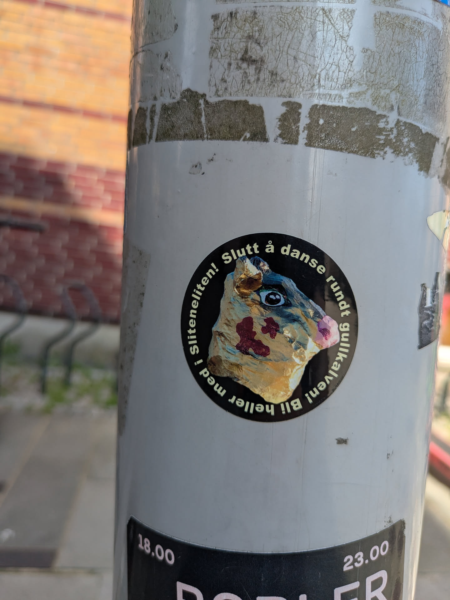 The sticker is circular with a black background. In the center is an image of a stylized animal head, possibly a pig or boar, with large, striking blue eyes, a pink snout, and reddish-brown patches on its face, giving it a somewhat abstract or deconstructed appearance. The creature's head is rendered in shades of gold, brown, and light blue. Around the central image, white text is arranged in a circular fashion. The sticker is affixed to a light grey pole, which shows signs of wear and tear, including peeling layers and dirt marks.
