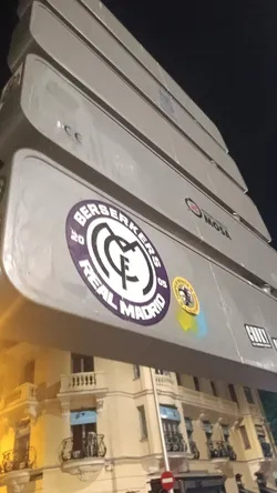 Street sticker The central sticker is a large, circular design featuring the Real Madrid Club de Fútbol logo. It has a prominent purple outer ring with white text that reads "BERSERKERS" across the top and "REAL MADRID" across the bottom. The numbers "20" and "05" are positioned on the left and right sides of the ring, respectively. The inner circle is white and contains the classic black and white intertwined "MCF" (Madrid Club de Fútbol) monogram, topped with a crown, which is the official emblem of Real Madrid. To the right of this main sticker, there is a smaller, circular yellow sticker. This smaller sticker displays a black and white portrait of a man, possibly a historical or political figure, surrounded by black text. The clearly readable text on this smaller sticker is "TIEMPO DE RESISTIR". Below this yellow sticker, there are faint blue and yellow spray paint marks.