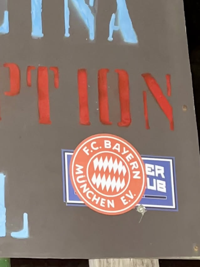 The sticker is a circular emblem of FC Bayern München. It features a vibrant red outer ring with white sans-serif text that reads "F.C. BAYERN MÜNCHEN E.V." arranged in an arc. The inner circle has a white background and displays a distinctive red and white diamond (lozenge) pattern, which is characteristic of the Bavarian flag. The overall design is clean, official, and instantly recognizable as the crest of the renowned German football club. It is affixed to a dark grey surface, partially overlapping a blue rectangular sticker with some white text visible underneath.