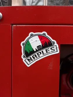 The sticker is an irregularly shaped design, resembling a cog or gear with rounded protrusions along its top and bottom edges, outlined in black and then white. The central part of the sticker features the vertical tricolor of the Italian flag: green on the left, white in the middle, and red on the right. Superimposed on this flag background is a black silhouette of a cityscape, depicting various buildings and a prominent historical structure, likely a landmark of Naples, situated towards the right side. Above the cityscape, within the white and red sections of the flag, are small black silhouettes of birds in flight. Below the cityscape, a white banner-like element curves upwards, displaying the word "NAPLES" in bold, capitalized, serif-style lettering with a subtle shadow effect. The sticker is applied to a textured, matte red surface.