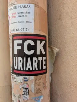 A sticker with the text "FCK URIARTE" in white on a black background with a red line on the top and bottom. The pole also has other stickers, including a pest control sticker that says "CONTROL DE PLAGAS, STOP cucarachas, STOP chinches, Económico - Rápido - Eficaz, (690 66 07 74".