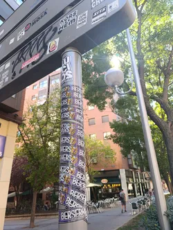 Street sticker The sticker is a long, continuous band wrapped around a metallic pole, creating a spiraling effect. It features a repeating design with the word 'BERSERKERS' prominently displayed in large, white, block letters with a black outline. Below 'BERSERKERS', the words 'Real Madrid' are written in a smaller, white, sans-serif font. To the left of the text, there is a stylized logo of an animal head, resembling a lion or tiger, in gold/yellow with black outlines, possibly wearing a crown, set within a shield or crest shape. The background of the sticker band is a vibrant purple. The sticker shows some signs of wear and slight peeling at the edges, indicating it's a physical application.