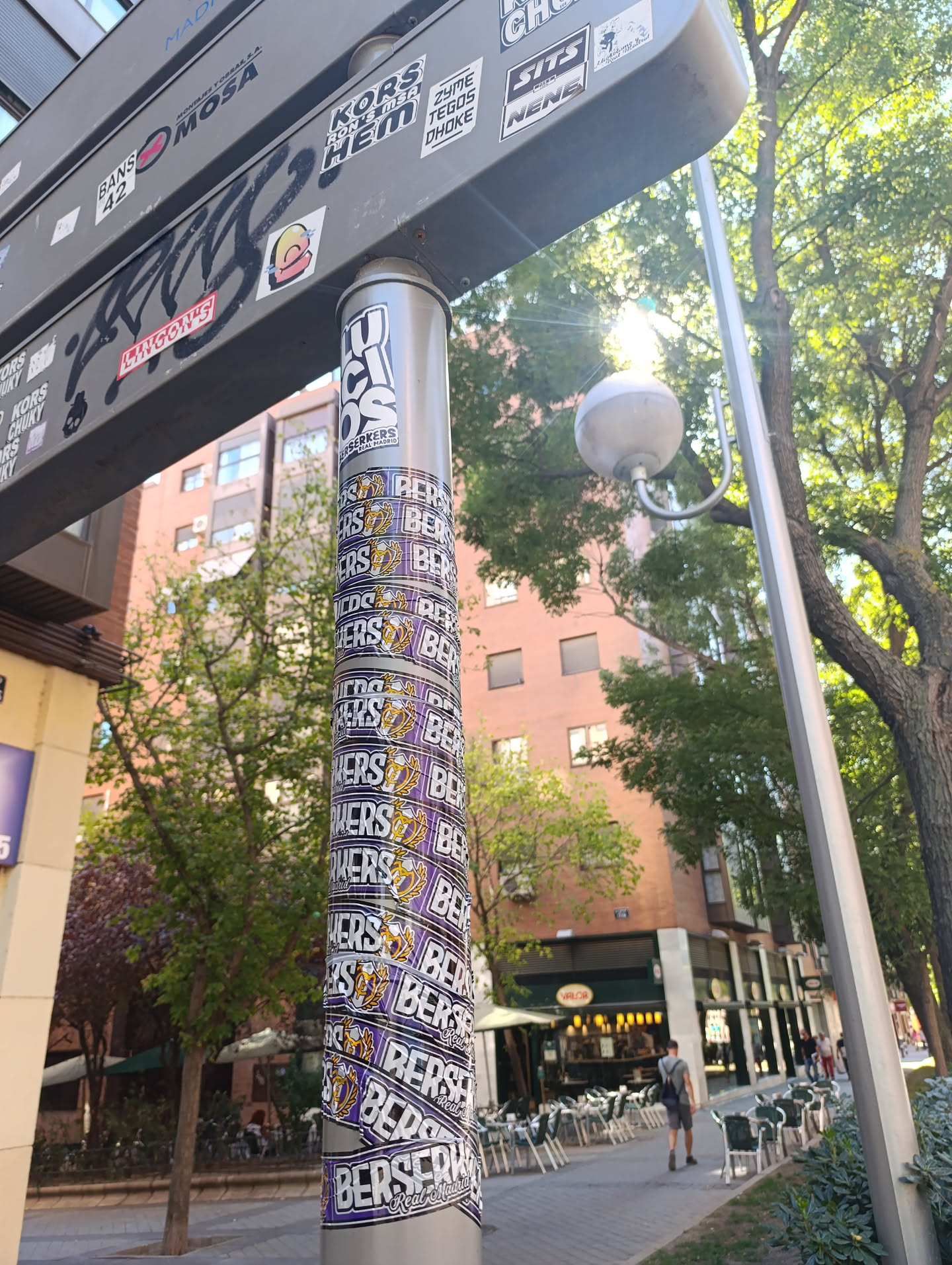 The sticker is a long, continuous band wrapped around a metallic pole, creating a spiraling effect. It features a repeating design with the word 'BERSERKERS' prominently displayed in large, white, block letters with a black outline. Below 'BERSERKERS', the words 'Real Madrid' are written in a smaller, white, sans-serif font. To the left of the text, there is a stylized logo of an animal head, resembling a lion or tiger, in gold/yellow with black outlines, possibly wearing a crown, set within a shield or crest shape. The background of the sticker band is a vibrant purple. The sticker shows some signs of wear and slight peeling at the edges, indicating it's a physical application.