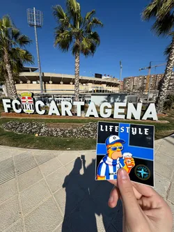 Street sticker A rectangular sticker featuring a stylized, yellow-skinned character resembling Homer Simpson, wearing white sunglasses, a blue and white striped baseball cap with a white 'S' logo, and a matching striped scarf. He is depicted holding a frothy glass of amber beer. The background is divided into black and blue sections. The word 'LIFESTYLE' is prominently displayed in white at the top. The word 'MALAGA' is visible on the character's scarf. In the bottom right corner, there is a circular logo with a white compass rose design and the text 'PERIANA ON TOUR' in white, set against a black background with a blue border.