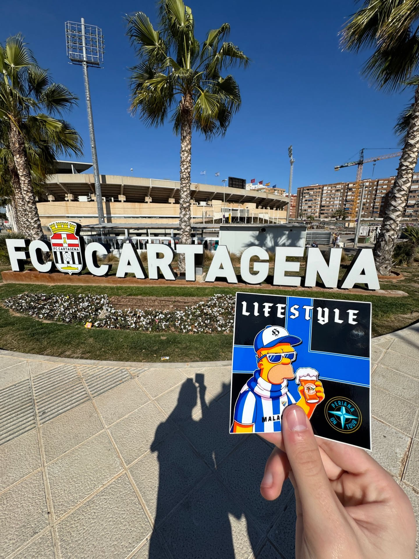 A rectangular sticker featuring a stylized, yellow-skinned character resembling Homer Simpson, wearing white sunglasses, a blue and white striped baseball cap with a white 'S' logo, and a matching striped scarf. He is depicted holding a frothy glass of amber beer. The background is divided into black and blue sections. The word 'LIFESTYLE' is prominently displayed in white at the top. The word 'MALAGA' is visible on the character's scarf. In the bottom right corner, there is a circular logo with a white compass rose design and the text 'PERIANA ON TOUR' in white, set against a black background with a blue border.