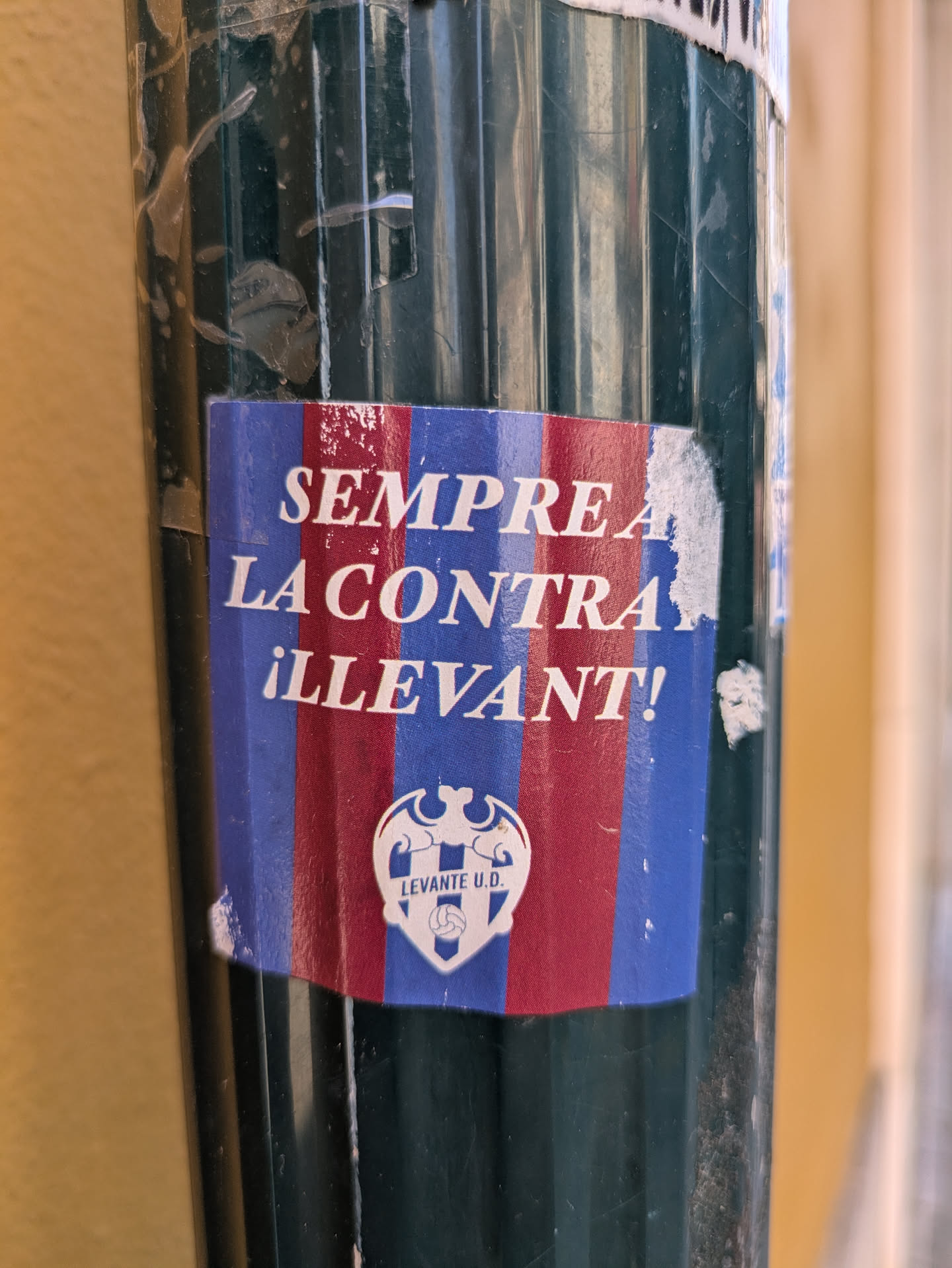 A weathered Levante UD sticker is affixed to a ribbed, dark green pole. The sticker features a bold, sans-serif typeface in white against a background of alternating vertical stripes in red and blue. The text reads "SEMPRE LA CONTRA ¡LLEVANT!" Beneath the text is the Levante UD crest. The sticker is partially peeling and shows signs of wear.
