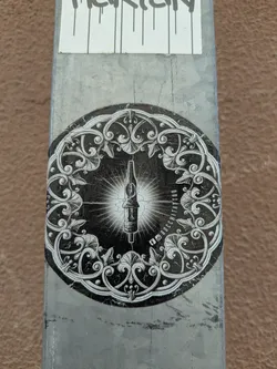 A round black and white sticker features a tattoo machine in the center, surrounded by an ornate, swirling floral design. Text around the right side indicates social media links.