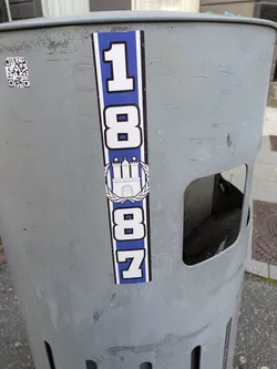 Street sticker The central sticker is vertical, predominantly blue and white. It displays the numbers '18' at the top, followed by a white crest of a castle with three towers (the Hamburg city crest) encircled by a wreath. Below the crest are the numbers '87'. The entire design is set against a blue background with a white border. The sticker is placed on a worn, light grey metal trash bin that has visible scratches and marks. To the left of the main sticker, a somewhat damaged QR code is visible.