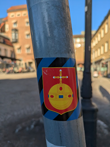 This sticker depicts a coat of arms, featuring a red shield with a yellow orb surmounted by a cross. The sticker is affixed to a metal pole and surrounded by alternating blue and black diagonal stripes.