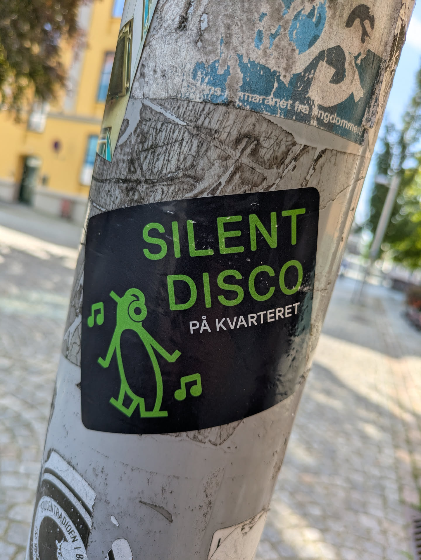 The sticker is rectangular with a black background and vibrant lime green text and graphics. The main text reads "SILENT DISCO" in large letters, followed by "PÅ KVARTERET" in smaller text below it. Below 'SILENT', there is a stylized green stick figure, possibly representing a person wearing headphones, with musical notes floating around its head and to its sides, indicating a dancing or music-related theme. The sticker is affixed to a weathered light pole covered with other faded and torn paper remnants.