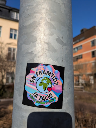 A square sticker with rounded corners on a metal pole. The main design features a colorful, marbled background framing a globe with a smiley face and a heart. The text reads "En Framtid? Ja Tack!" and "Rösta Grönt 9 Juni" suggesting a call to vote green. It is an environmental advocacy.