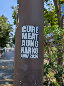 Street sticker A rectangular black sticker with bold white text reading 'CURE MEAT AUNG HARKO AOW 2026' stacked vertically, affixed to a brown pole.