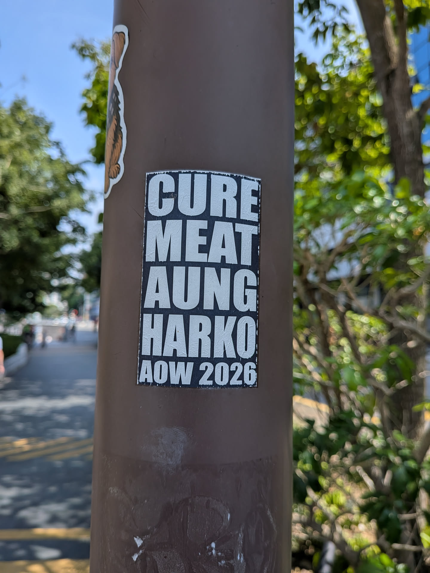 A rectangular black sticker with bold white text reading 'CURE MEAT AUNG HARKO AOW 2026' stacked vertically, affixed to a brown pole.