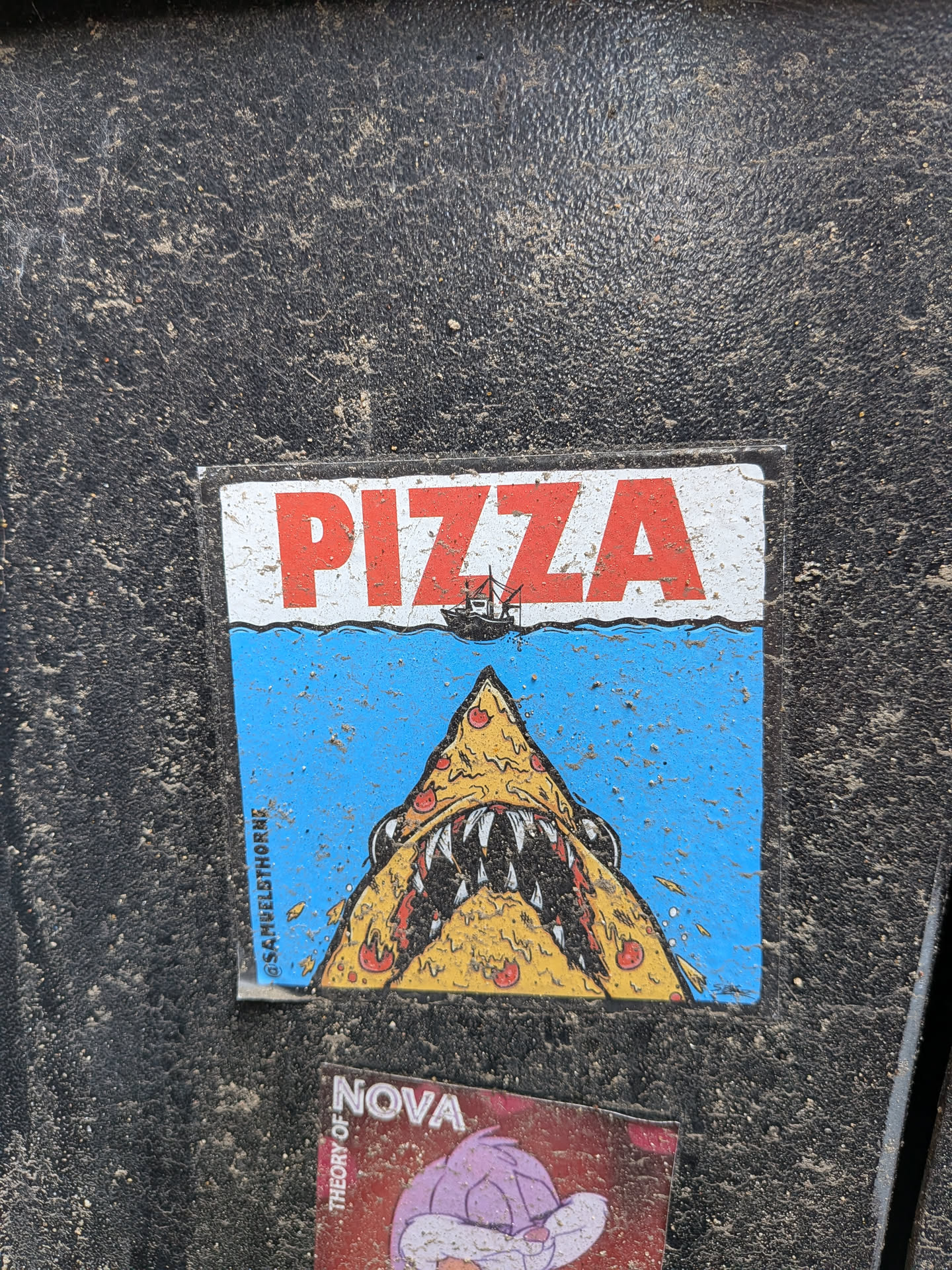 A rectangular sticker featuring a humorous illustration. The top portion is white with the word "PIZZA" in large, bold red letters. Below this, a wavy line separates the white background from a vibrant blue ocean scene. A small, dark fishing boat with a red sail is depicted on the horizon, positioned under the 'ZZA' of 'PIZZA'. Emerging from the blue water is a large, menacing pizza slice, designed to resemble a shark's head with sharp white teeth and dark eyes. The pizza slice is yellow with red pepperoni toppings and dark crust lines. Small splashes or debris are shown around the pizza shark. Along the left edge, oriented vertically, is a social media handle. The sticker appears slightly worn and is covered with specks of dirt, indicating it's affixed to an outdoor surface.