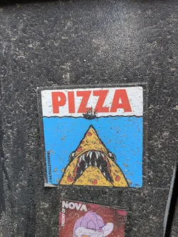 Street sticker A rectangular sticker featuring a humorous illustration. The top portion is white with the word "PIZZA" in large, bold red letters. Below this, a wavy line separates the white background from a vibrant blue ocean scene. A small, dark fishing boat with a red sail is depicted on the horizon, positioned under the 'ZZA' of 'PIZZA'. Emerging from the blue water is a large, menacing pizza slice, designed to resemble a shark's head with sharp white teeth and dark eyes. The pizza slice is yellow with red pepperoni toppings and dark crust lines. Small splashes or debris are shown around the pizza shark. Along the left edge, oriented vertically, is a social media handle. The sticker appears slightly worn and is covered with specks of dirt, indicating it's affixed to an outdoor surface.