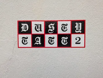 Street sticker A rectangular sticker with a thin red border containing a 2x5 checkerboard grid of alternating black and white squares. Each square features a single Gothic-style letter or number, spelling 'DUSTY' on the top row and 'TATT2' on the bottom row.