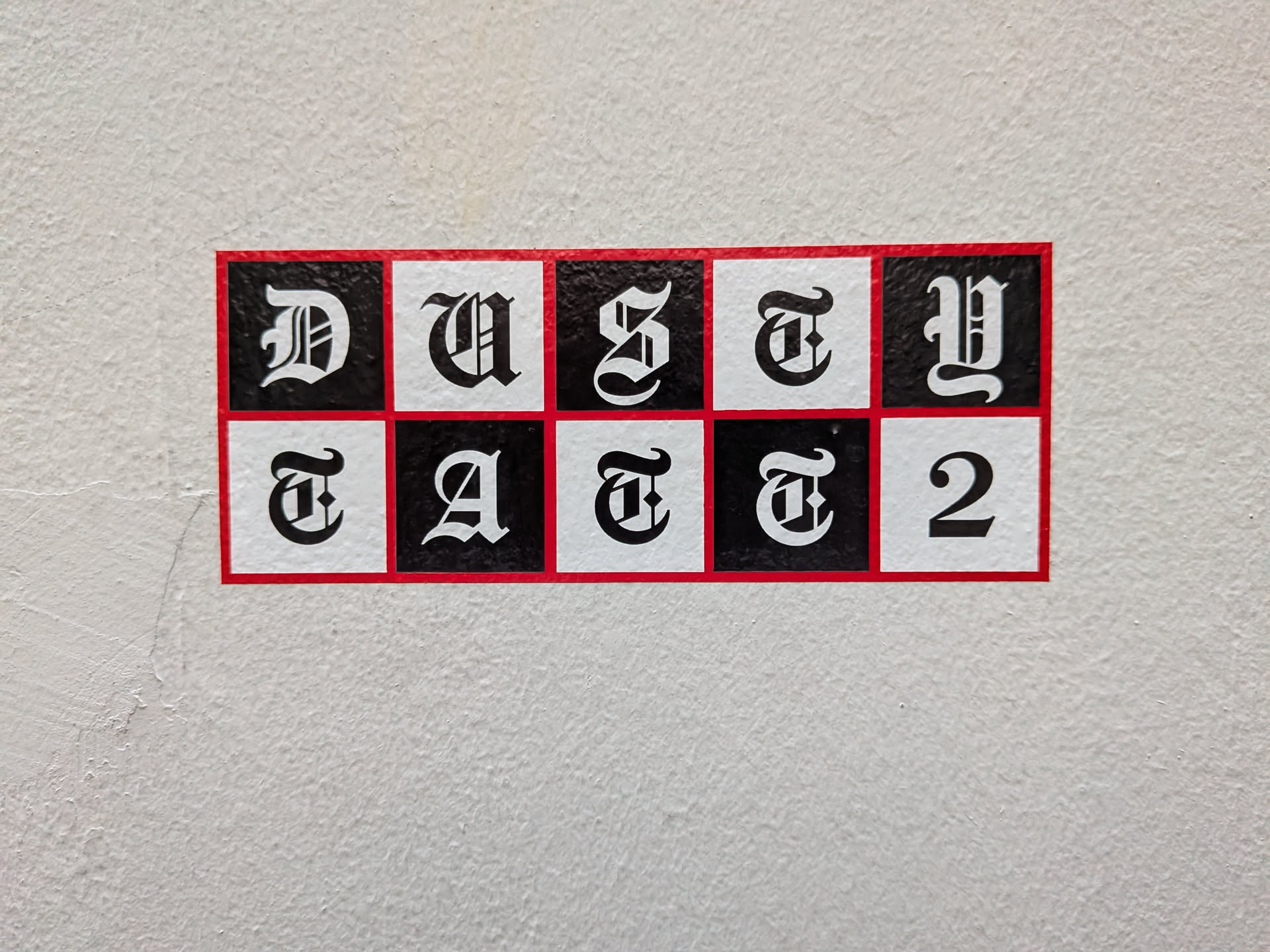 A rectangular sticker with a thin red border containing a 2x5 checkerboard grid of alternating black and white squares. Each square features a single Gothic-style letter or number, spelling 'DUSTY' on the top row and 'TATT2' on the bottom row.