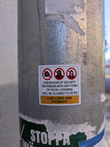 The sticker on the pole features three images with red circles and a line through them, depicting various forms of facial coverings that are not allowed. The text reads: "FOR REASON OF SECURITY NO PERSON WITH ANY FORM OF FACIAL COVERING WILL BE ALLOWED TO ENTER LIVE LONG AND PROSPER".