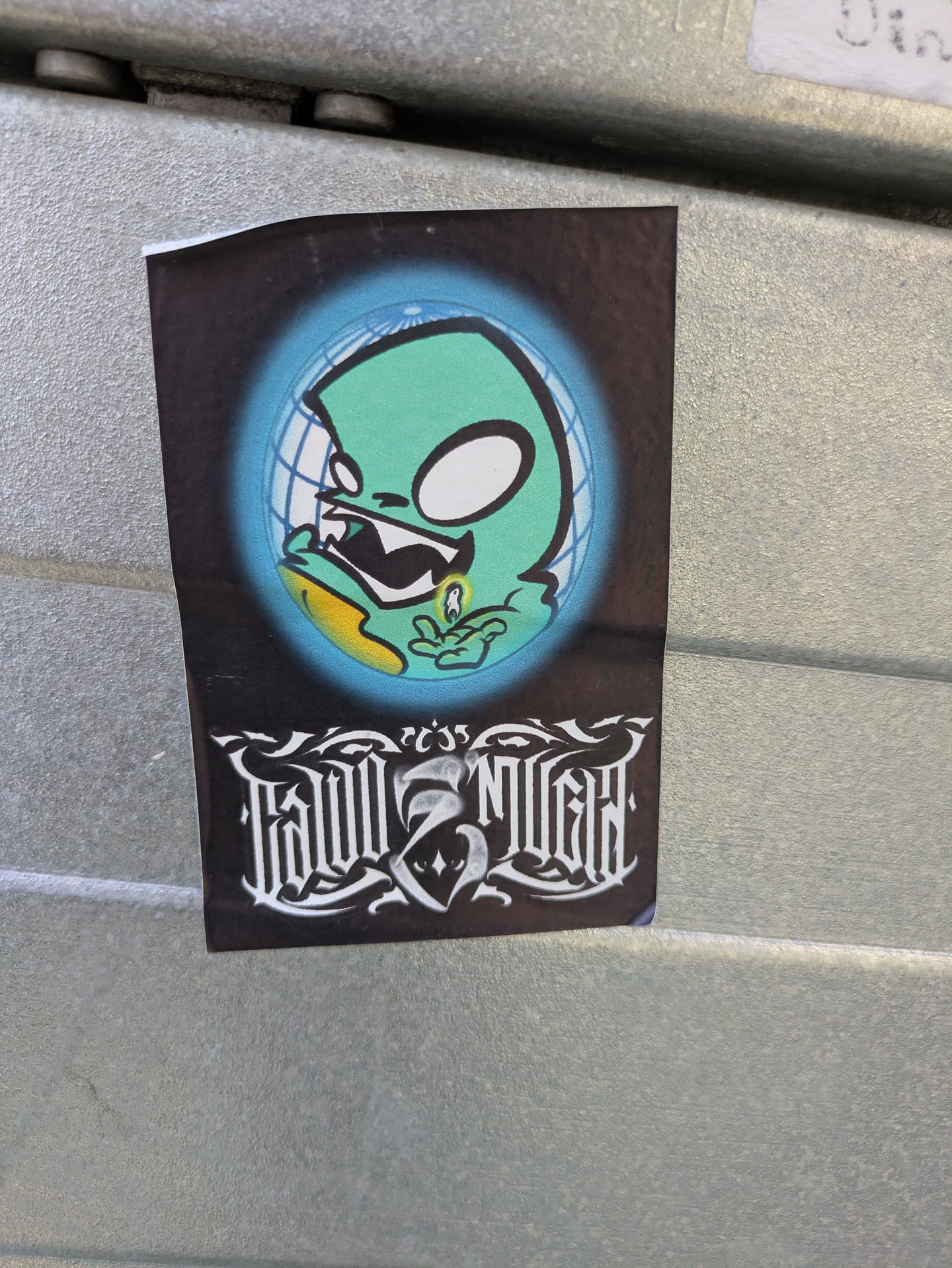 The sticker displays a stylized, light green alien or monster character with large white, expressive eyes, an open mouth showing sharp teeth, and what appears to be drool emanating from its mouth. It is depicted inside a spherical, blue glowing outline, resembling a globe with a grid pattern. The background of the sticker is dark. Below the main image, there is intricate, white gothic-style lettering. Above the second word, there are small numbers '235'. The sticker is applied to a textured, grey metallic surface.