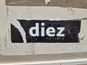 A black and white sticker with the word "diez" in a bold, sans-serif font. Below it, in a smaller font, it says "pizza co". The sticker is slightly torn at the edges and shows signs of wear. It is adhered to a wall that has other markings and discoloration.