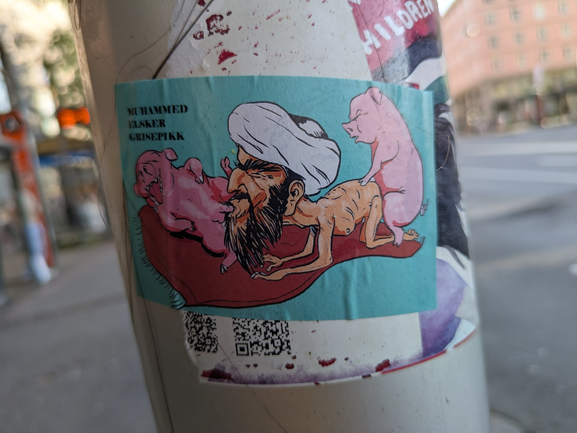 The sticker features a cartoon illustration of a bearded man wearing a white turban, widely recognized as a caricature of Prophet Muhammad, lying on a red mat or cushion. Two pink pigs are interacting with him: one appears to be nuzzling or licking his face, while the other is positioned suggestively behind him. In the upper left corner, the text "MUHAMMED ELSKER GRISEPIKK" is written, which translates from Norwegian to "MUHAMMED LOVES PIG PENIS," a phrase deliberately chosen for its extreme blasphemous and offensive nature within an Islamic context. The sticker is affixed to a pole in a public urban setting, aimed at provoking strong reactions and is indicative of anti-religious or islamophobic sentiment.