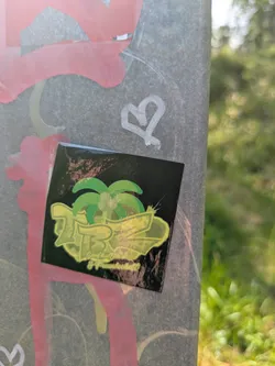 Street sticker The sticker is square with a glossy finish and a black background. The central design includes a vibrant green palm tree. Overlapping and in front of the palm tree is large, stylized text that appears to read 'ZB', rendered in a bright yellow-green outline with a darker green fill, giving it a layered, almost three-dimensional effect. Beneath this large lettering, in a smaller, lighter font, the German words 'Gaufkultur & Freundschaft' are visible. The sticker is placed on a textured grey pole or wall, which also bears pink and white graffiti marks, including a heart symbol.