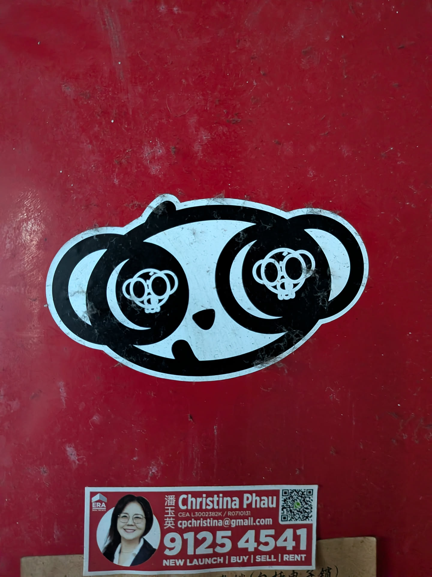 A black and white die-cut sticker of a stylized panda face. The eyes contain small skull-like graphics. It is placed on a red surface.