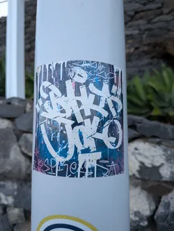 A rectangular sticker featuring bold, white graffiti-style lettering over a textured blue and pink background with paint drips. Smaller stylized tags are visible along the top and bottom borders.