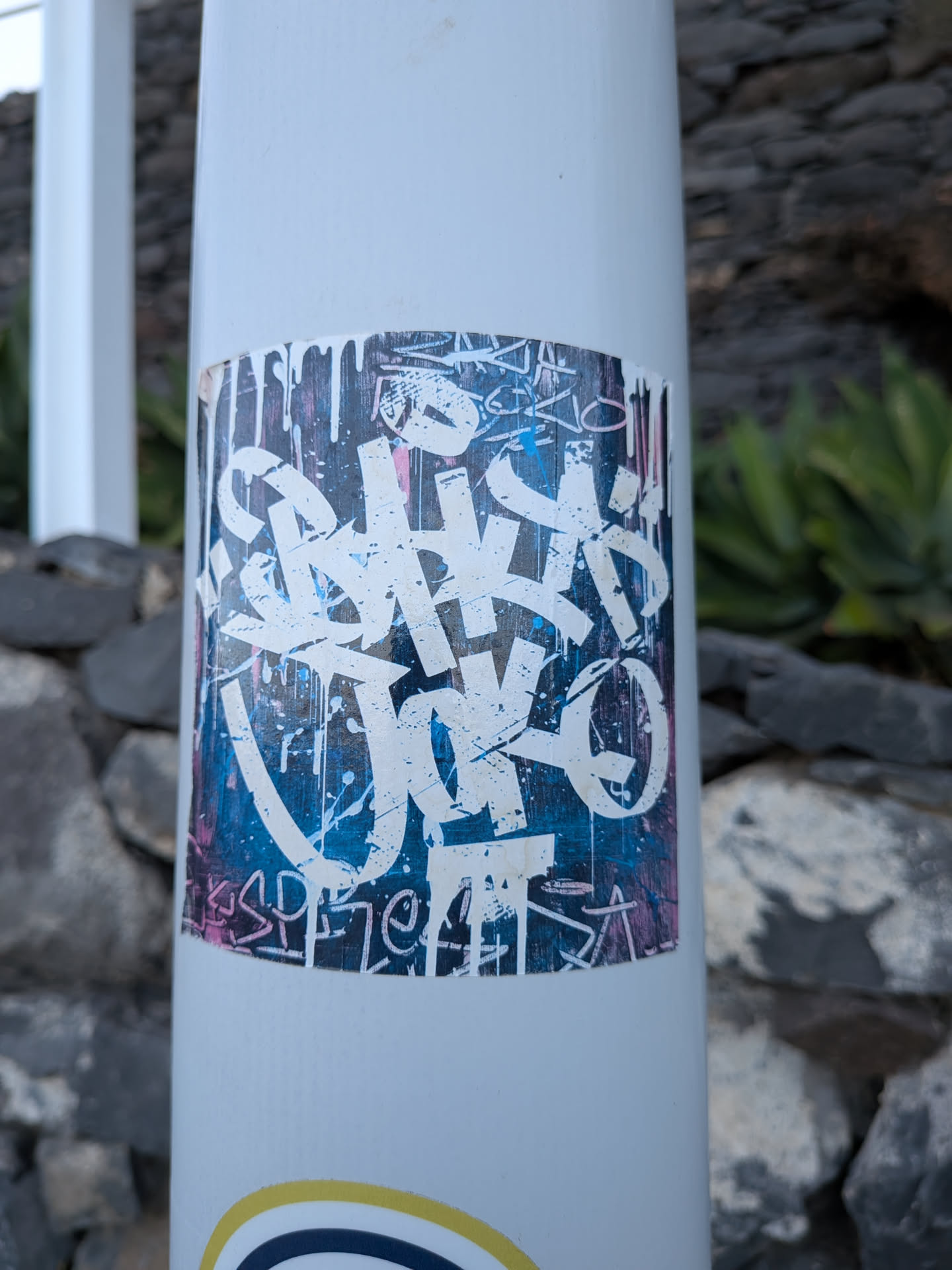 A rectangular sticker featuring bold, white graffiti-style lettering over a textured blue and pink background with paint drips. Smaller stylized tags are visible along the top and bottom borders.
