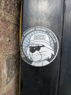 Street sticker A circular sticker featuring a cartoon cat's head and upper torso. The cat is depicted in a grayscale palette with white fur and black outlines. It wears dark, rectangular sunglasses and a white bucket hat with the letters 'ADFS' prominently displayed on the front. The background behind the cat consists of radiating light grey lines, creating a sunburst effect. A dark grey outer ring encircles the central design, containing white text. The sticker has a slightly worn appearance, indicating it's a physical object adhered to a surface.