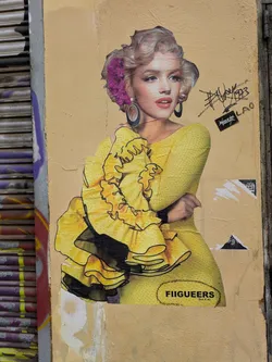 Street sticker This is a sticker featuring a modified image of Marilyn Monroe wearing a yellow dress with ruffles and a purple flower in her hair. The sticker is on a yellow wall. At the bottom, there's the text "FIIGUEERS" followed by "Rich & Art". Above the sticker, there are a few signatures and other small stickers.