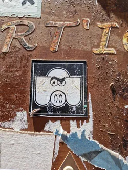 Street sticker A square sticker featuring a simple cartoon illustration of a character with angry eyebrows and a small mouth. The sticker has a black background with a white character in the center and a number 00 at the bottom. The sticker is pasted on a rusty brown wall covered in other stickers and graffiti.