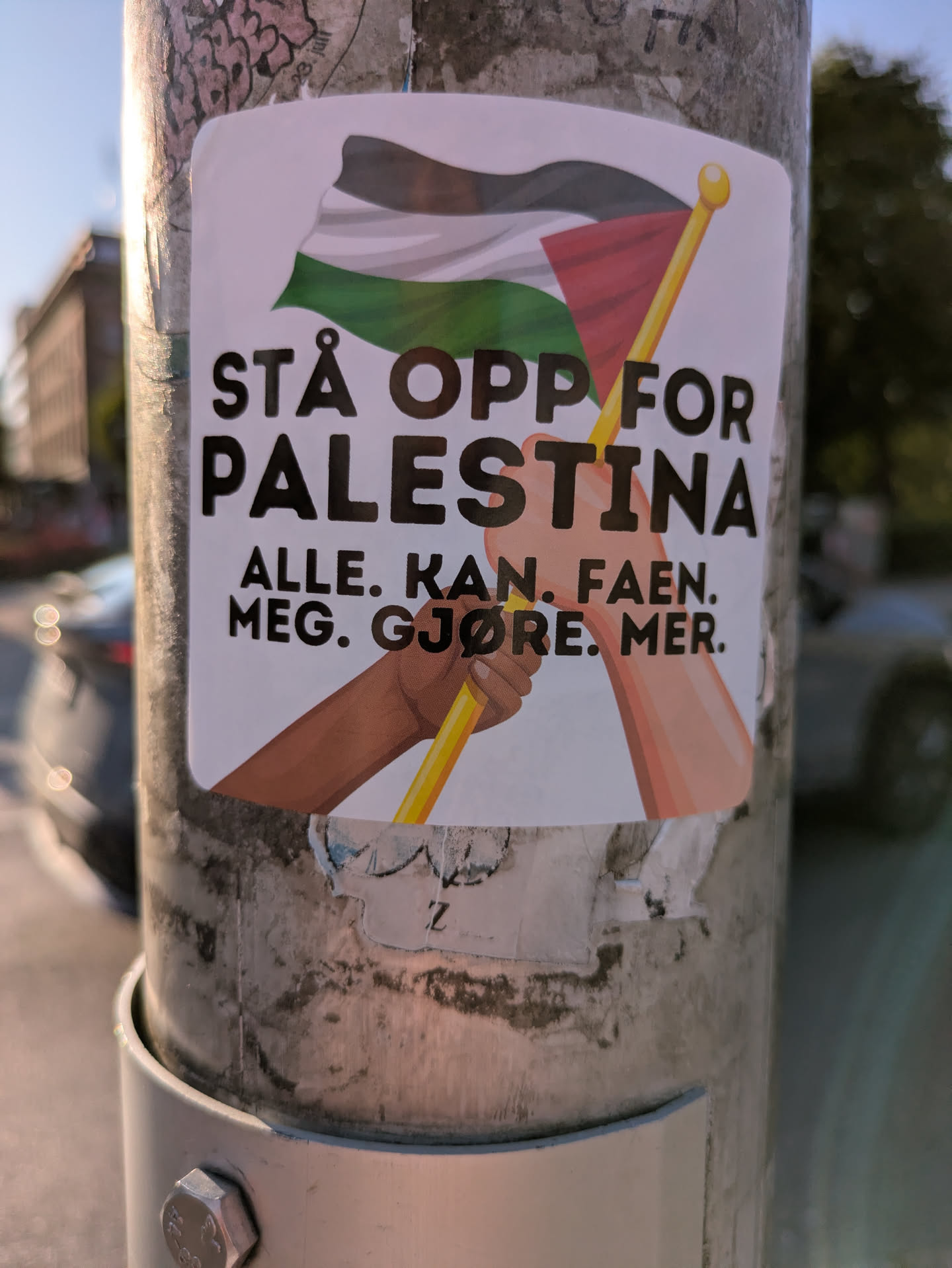 The sticker prominently displays the Palestinian flag waving at the top. Below the flag, two hands, one with a darker skin tone and the other lighter, are shown grasping the yellow flagpole together, symbolizing unity and shared effort. The text on the sticker is in Norwegian, set in a clear, bold, dark font against a white background. The sticker is affixed to a grey metal pole, which shows signs of wear and some faint markings from other removed adhesives. The background is blurred, suggesting an urban outdoor setting.