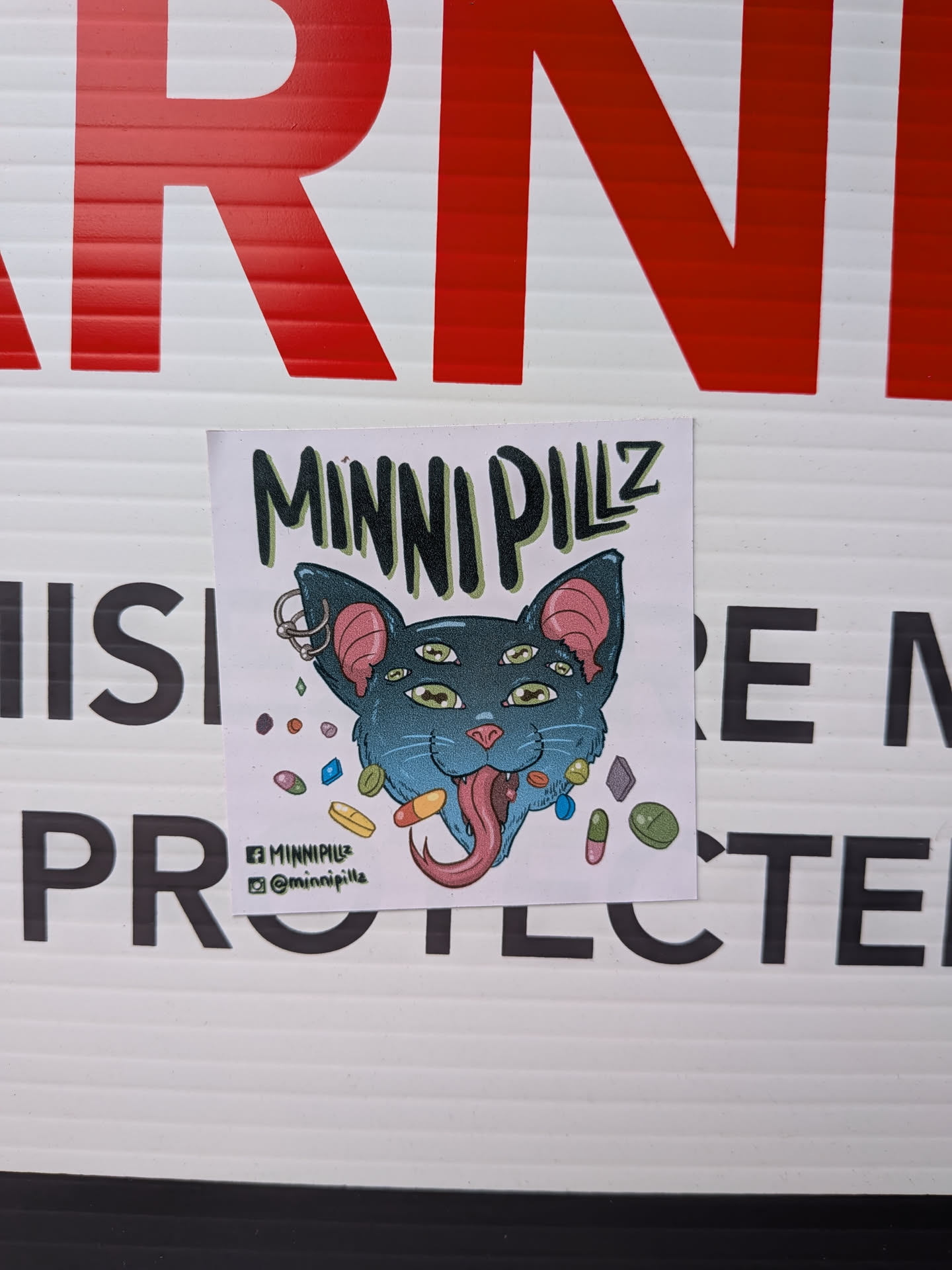 A square sticker featuring a stylized, cartoonish cat head. The cat is a shade of teal or blue-green with prominent pink inner ears and a pink nose. It has six yellow-green eyes with black pupils, giving it a psychedelic appearance. A long, pink, drooling tongue hangs from its open mouth. The cat's left ear is adorned with two silver hoop piercings. Scattered around the cat's head are various colorful pills and capsules in shades of purple, orange, green, blue, and yellow. Above the cat, the text "MINNI PILLZ" is written in a bold, dark green, somewhat jagged font. Below the cat, social media icons for Facebook and Instagram are present with corresponding handles. The background of the sticker is white.