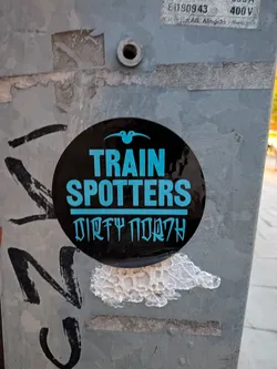 Street sticker The sticker is circular with a black background. At the top, a small, stylized blue bird with its wings spread is depicted. Below the bird, the words 'TRAIN SPOTTERS' are written in large, bold, light blue capital letters. Beneath this, a line of text in a stylized, somewhat runic font, also in light blue, reads 'DIRTY NORTH'. The sticker appears to be applied to a metal pole, with some surrounding graffiti and wear.