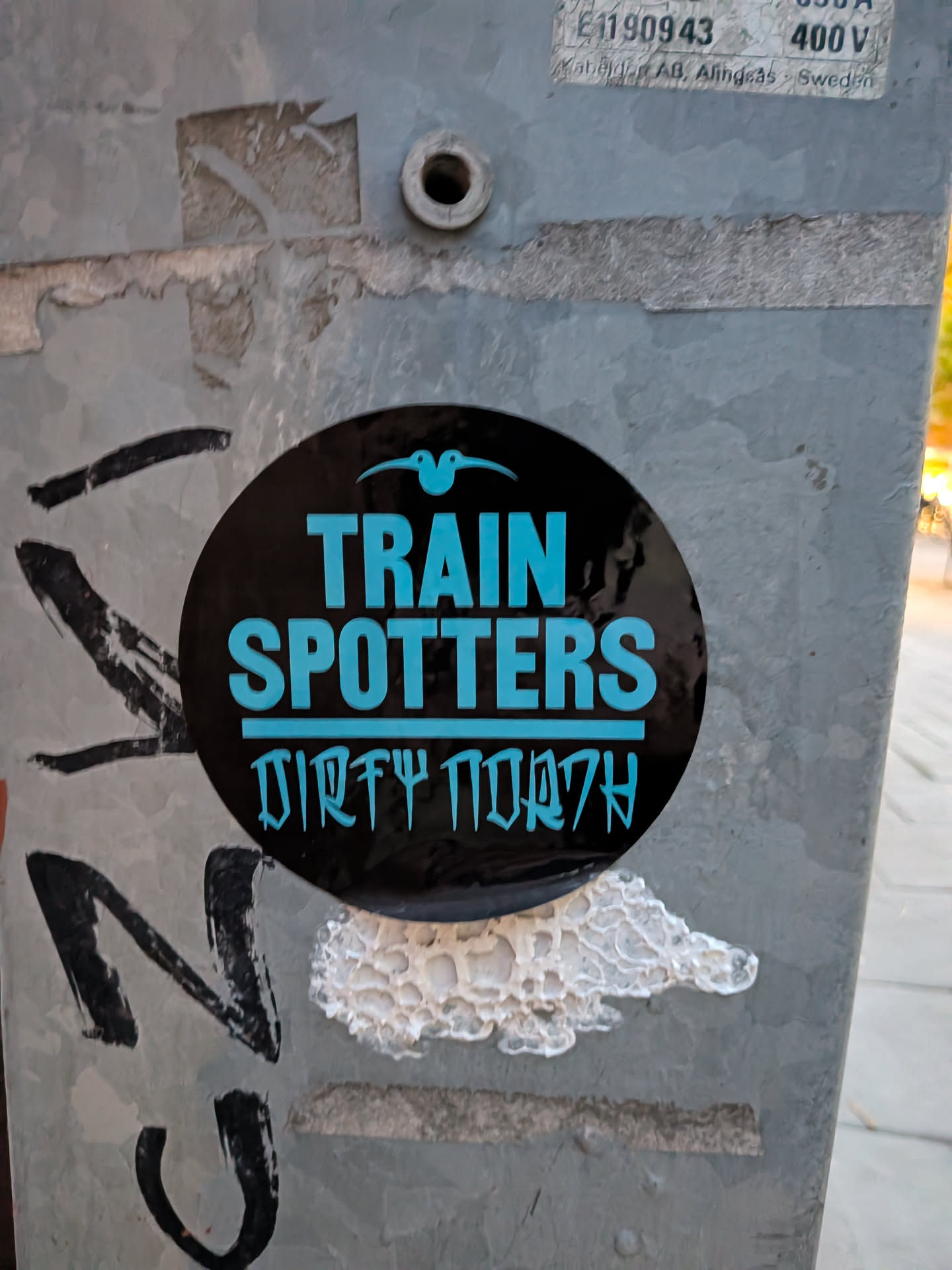 The sticker is circular with a black background. At the top, a small, stylized blue bird with its wings spread is depicted. Below the bird, the words 'TRAIN SPOTTERS' are written in large, bold, light blue capital letters. Beneath this, a line of text in a stylized, somewhat runic font, also in light blue, reads 'DIRTY NORTH'. The sticker appears to be applied to a metal pole, with some surrounding graffiti and wear.