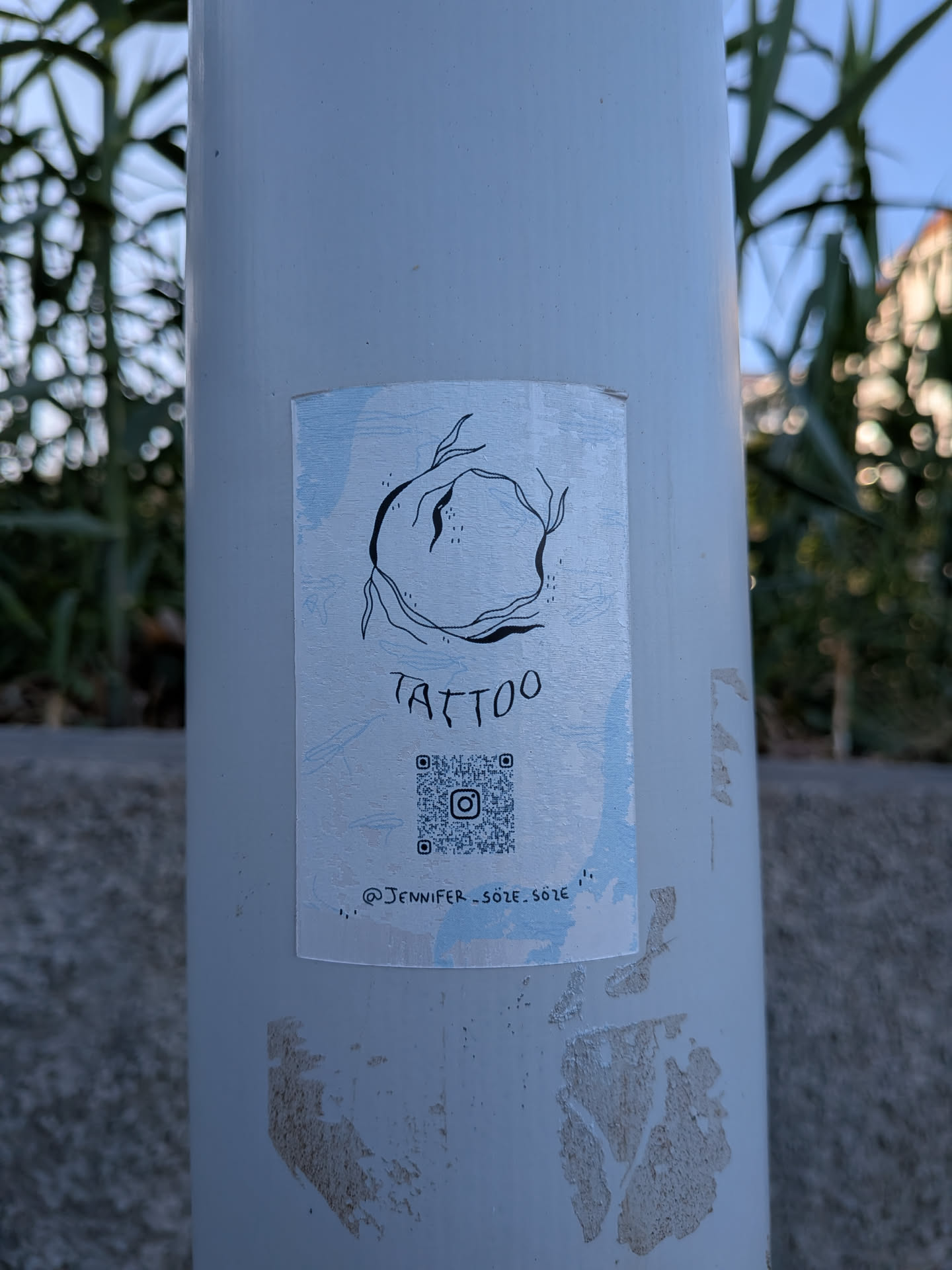 A rectangular sticker on a white pole with a light blue textured background. It displays a black line-art abstract circular design, the hand-drawn word 'TATTOO', a QR code with an Instagram logo, and the handle '@JENNIFER_SOZE_SOZE'.