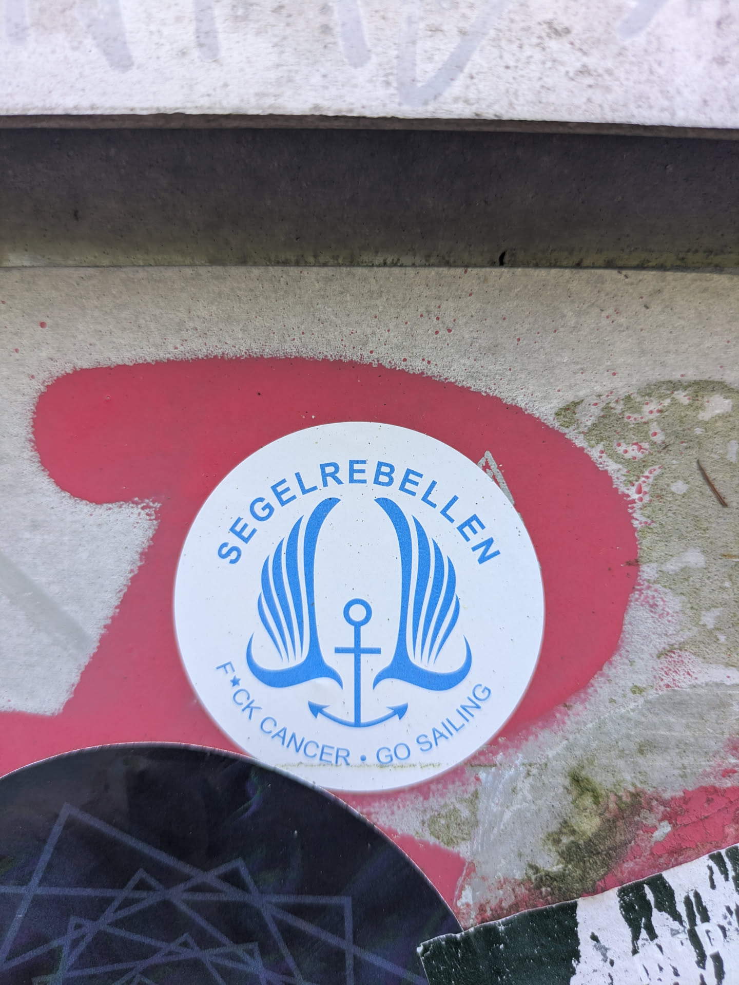The central image is a white circular sticker with a distinct blue design. It features a stylized anchor at its base, from which two large, flowing wing-like shapes extend upwards and outwards, giving the impression of movement or flight. Above this emblem, arcing along the top edge of the circle, is the word 'SEGELREBELLEN' in bold blue capital letters. Along the bottom edge, also in blue capital letters, is the defiant phrase 'F*CK CANCER • GO SAILING', with a small dot separating 'CANCER' and 'GO SAILING'. The sticker is applied to a gritty, light-colored surface, possibly concrete or a wall, which has remnants of bright red spray paint underneath and around it. Parts of other stickers are visible at the very bottom edge of the image, suggesting a location where multiple stickers have been placed.