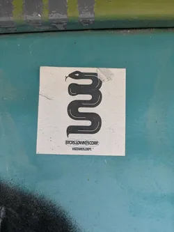 This is a white square sticker on a teal surface. The sticker features a black snake symbol that looks like stylized letter "M". Below the snake is text that reads "BTCRS.LOWKEY.CORP." and "WIZZARDS.DEPT."