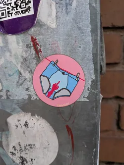Street sticker A round pink sticker depicting light blue panties hanging on a clothesline. The crotch area features a red bloodstain shaped like a hand giving the middle finger. Small yellow text at the bottom reads 'TIINA SÖÖT'.