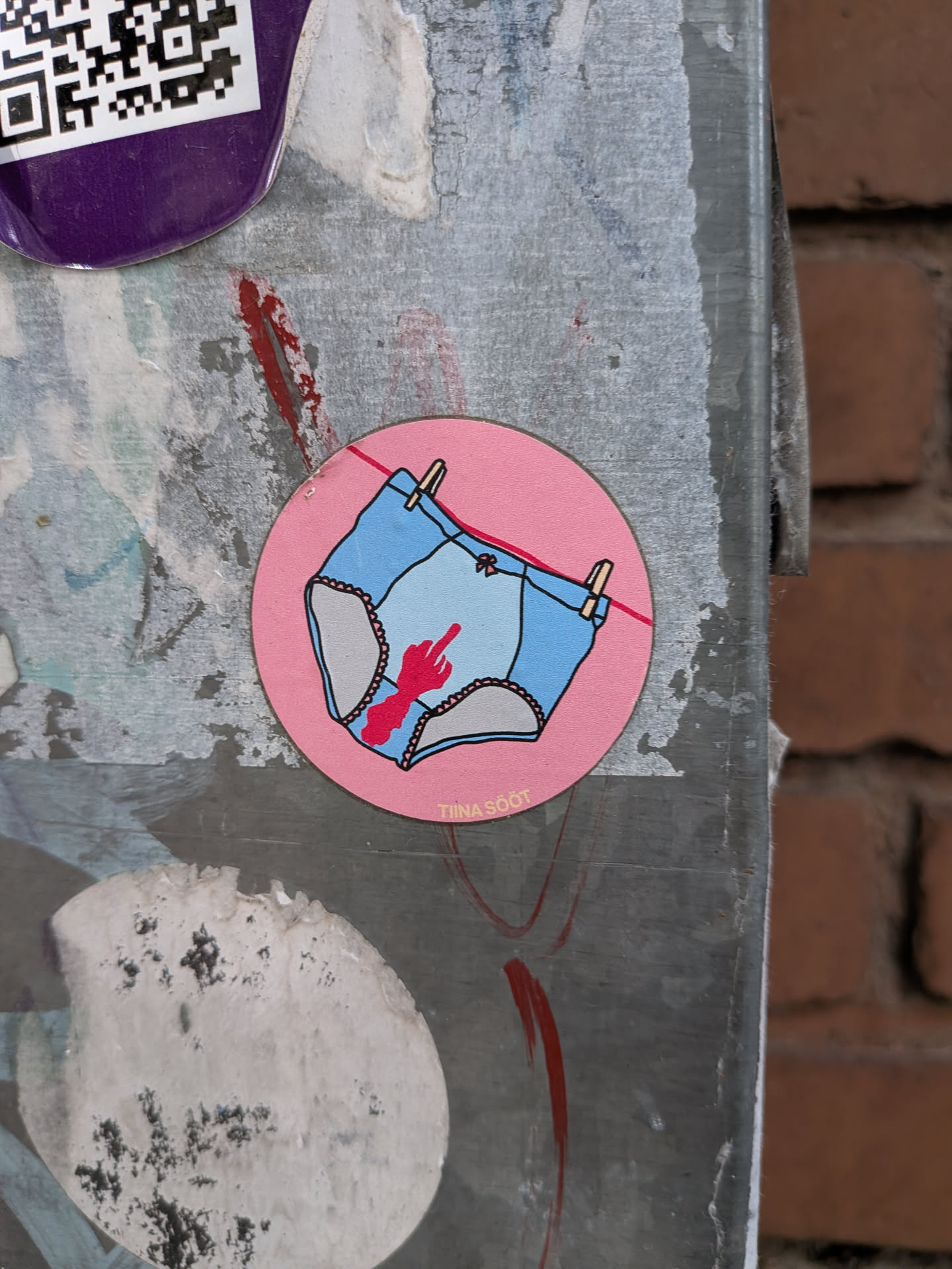 A round pink sticker depicting light blue panties hanging on a clothesline. The crotch area features a red bloodstain shaped like a hand giving the middle finger. Small yellow text at the bottom reads 'TIINA SÖÖT'.